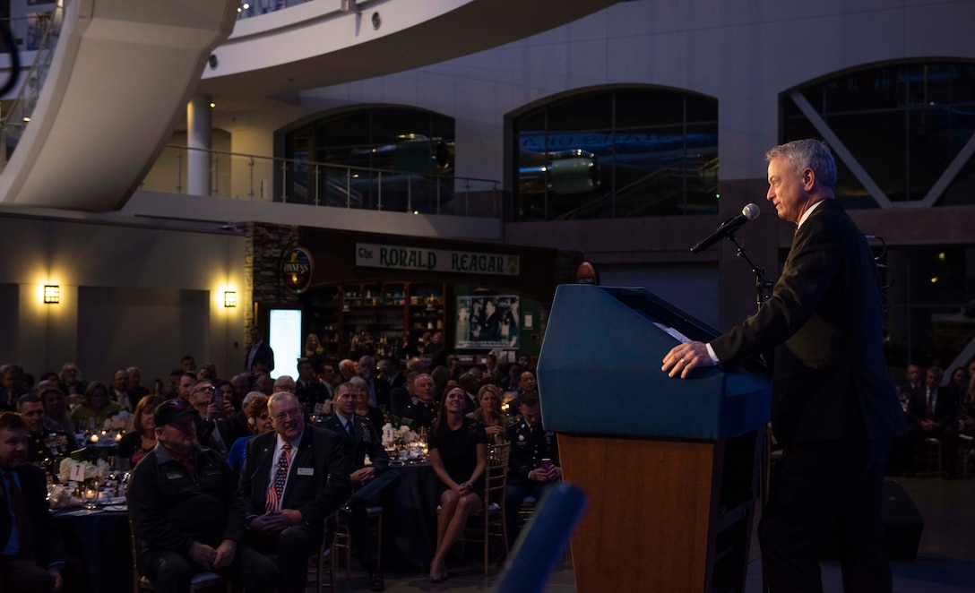 Man speaks at lectern as an audience seated at round tables listens.