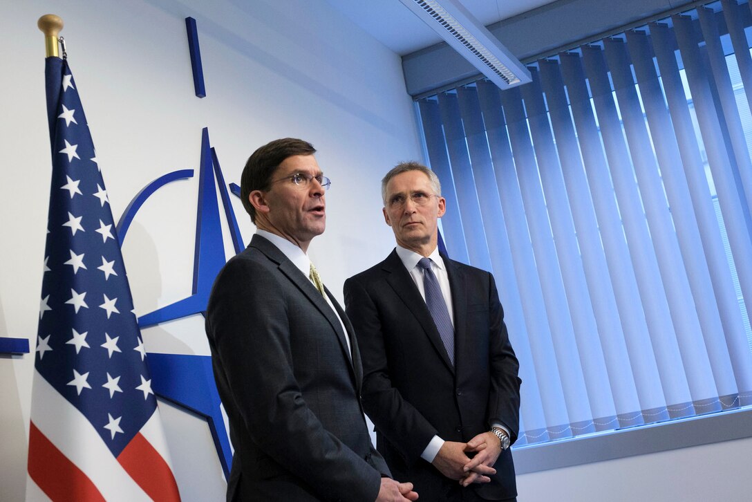 Defense Secretary Dr. Mark T. Esper speaks while standing with another official in front of a wall bearing the NATO logo.