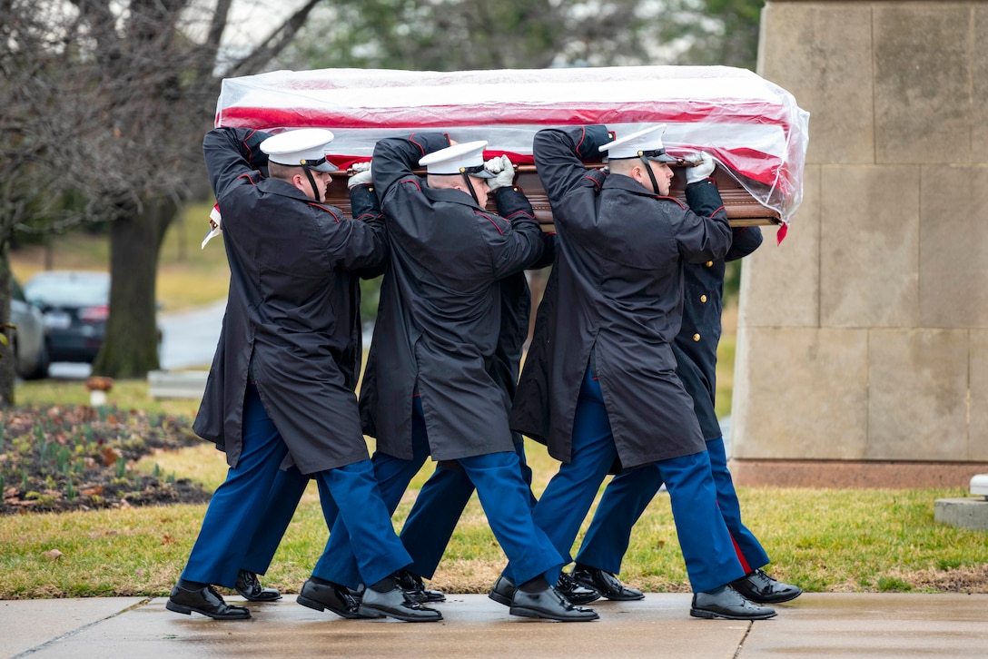 Soldiers and Marines walk in formation carrying a casket at a cemetery.