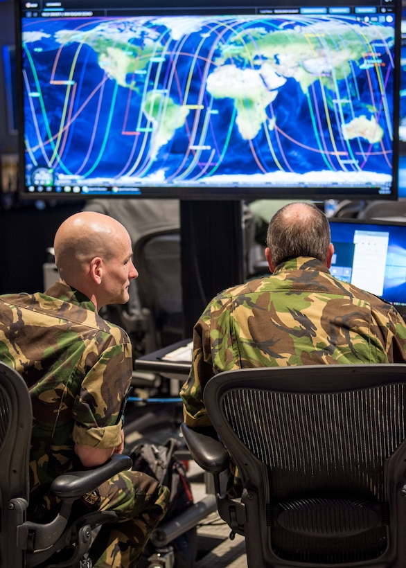 Two men in military uniforms sit looking at a monitor displaying a world map.