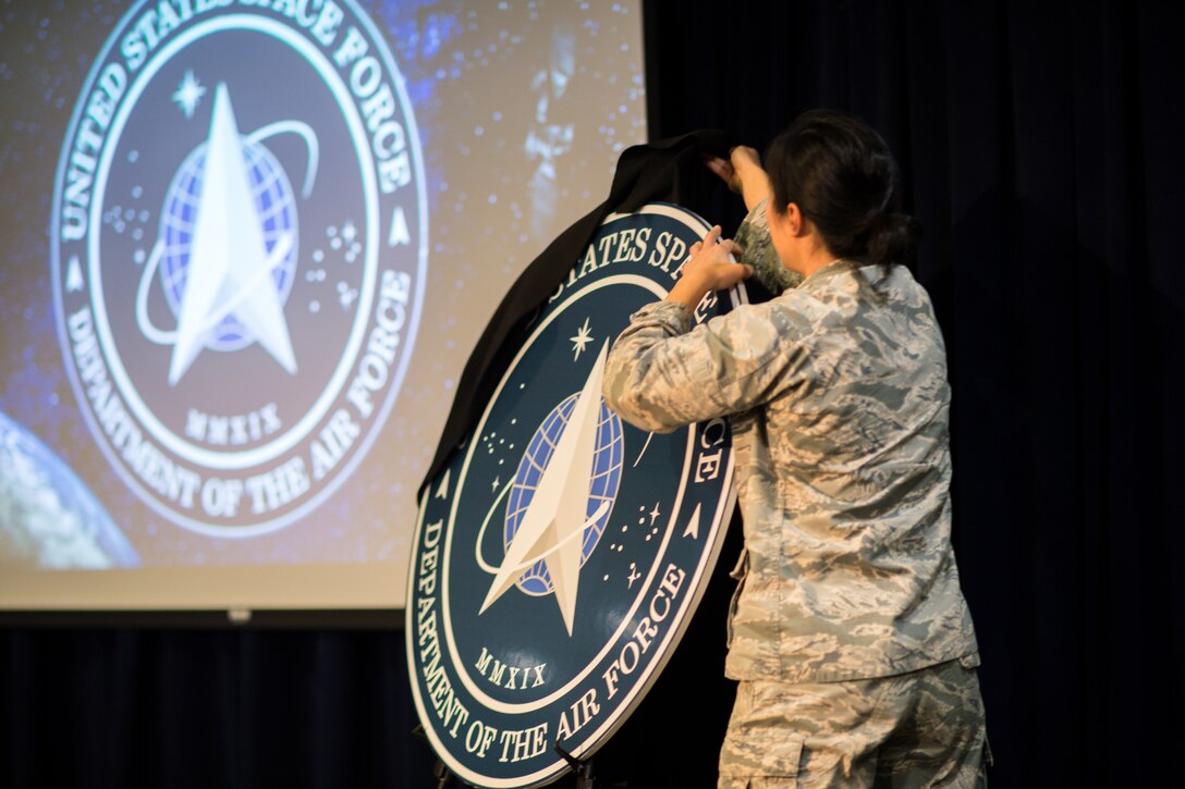 A uniformed service member pulls a black sheet off an easel that holds a circular cutout featuring the seal of the U.S. Space Force.