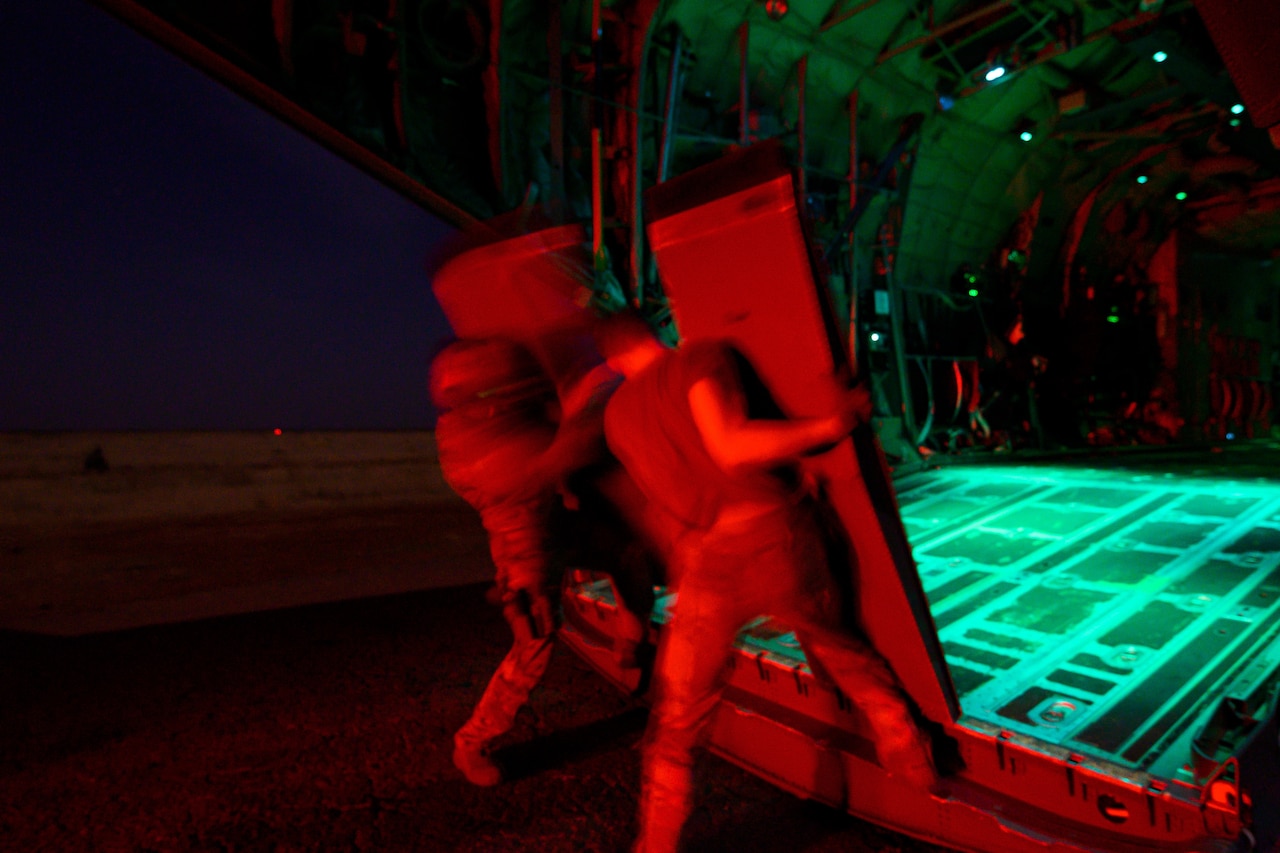 Two airmen, illuminated in red light, remove ramps from the back of an open aircraft in the dark.