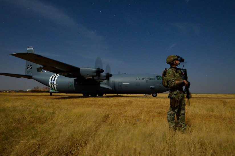 A soldier with a weapon stands by an aircraft in a field.