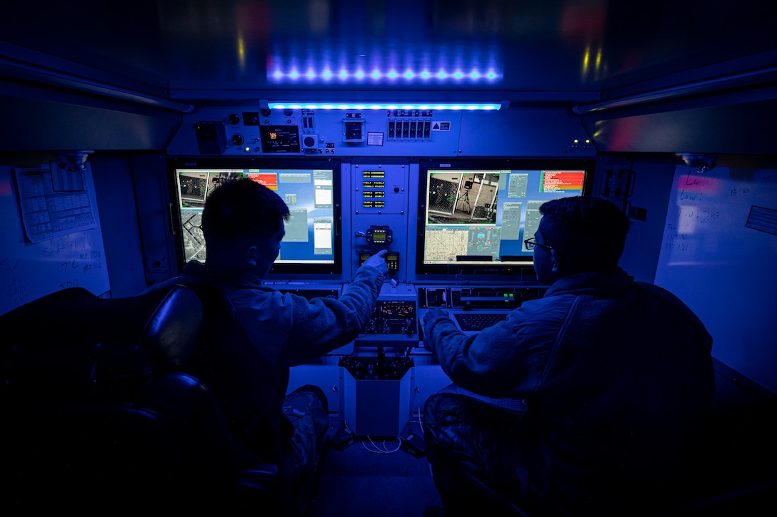 Soldiers sit in front of computer screens in an area illuminated by blue light.