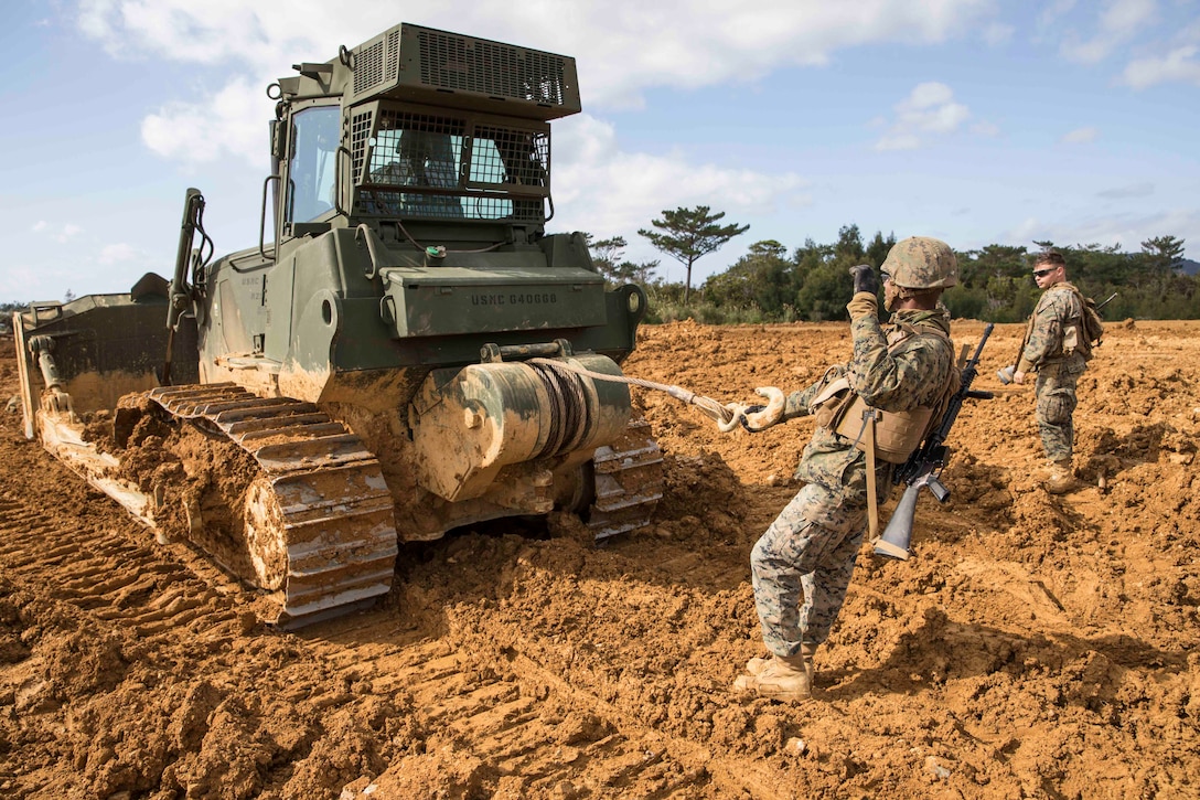 A Marine does a hand signal from behind heavy machinery as another Marine stands by.