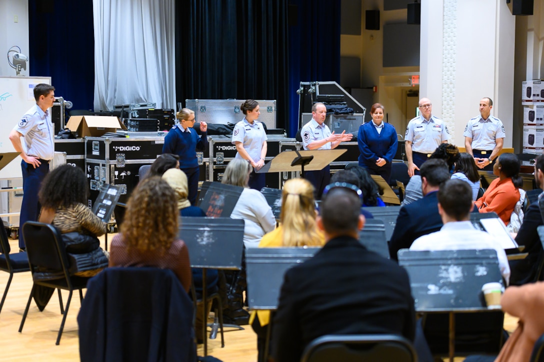 Seven people in Air Force duty blue uniform are standing in front of a crowd of 55 seated people dressed in civilian attire. One Air Force member in the center is gesturing and speaking while the other six Air Force members are looking at him.