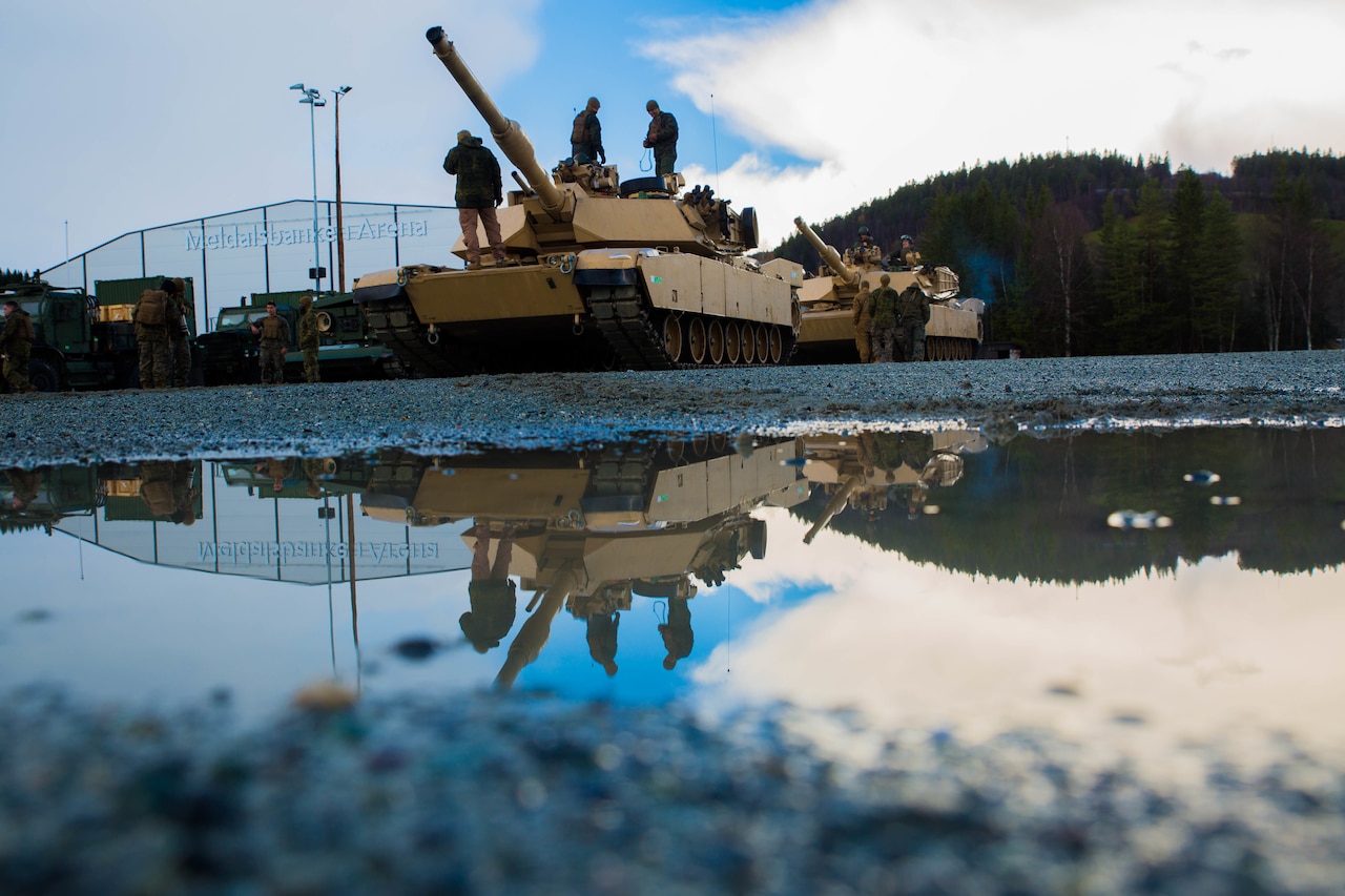 Service members stand atop a tank in a waterlogged dirt parking lot.