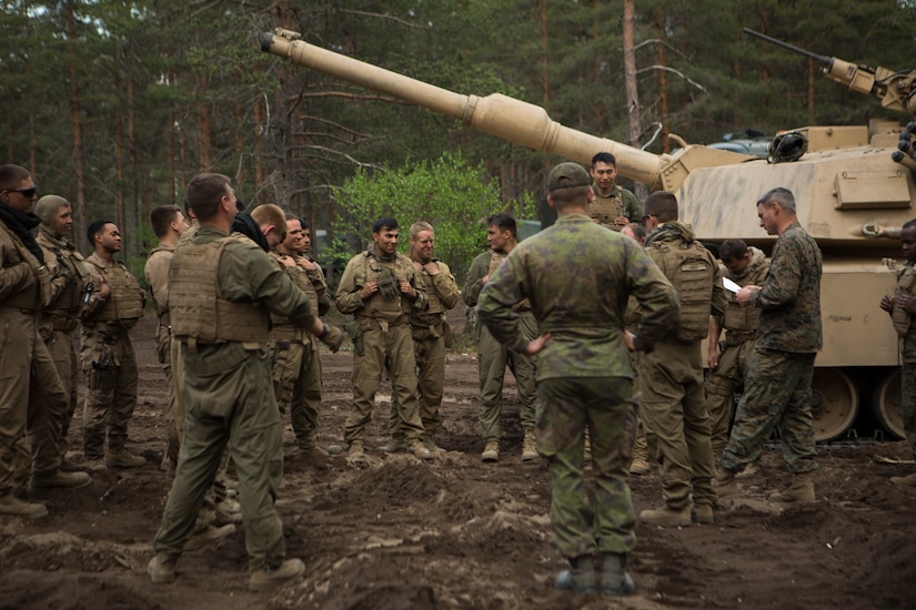 Service members stand near a tank in a forested area.