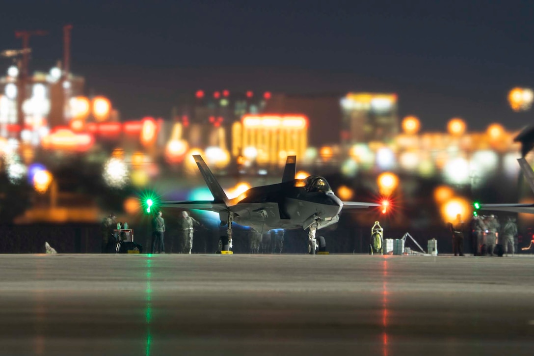Service members stand around a military jet at night.