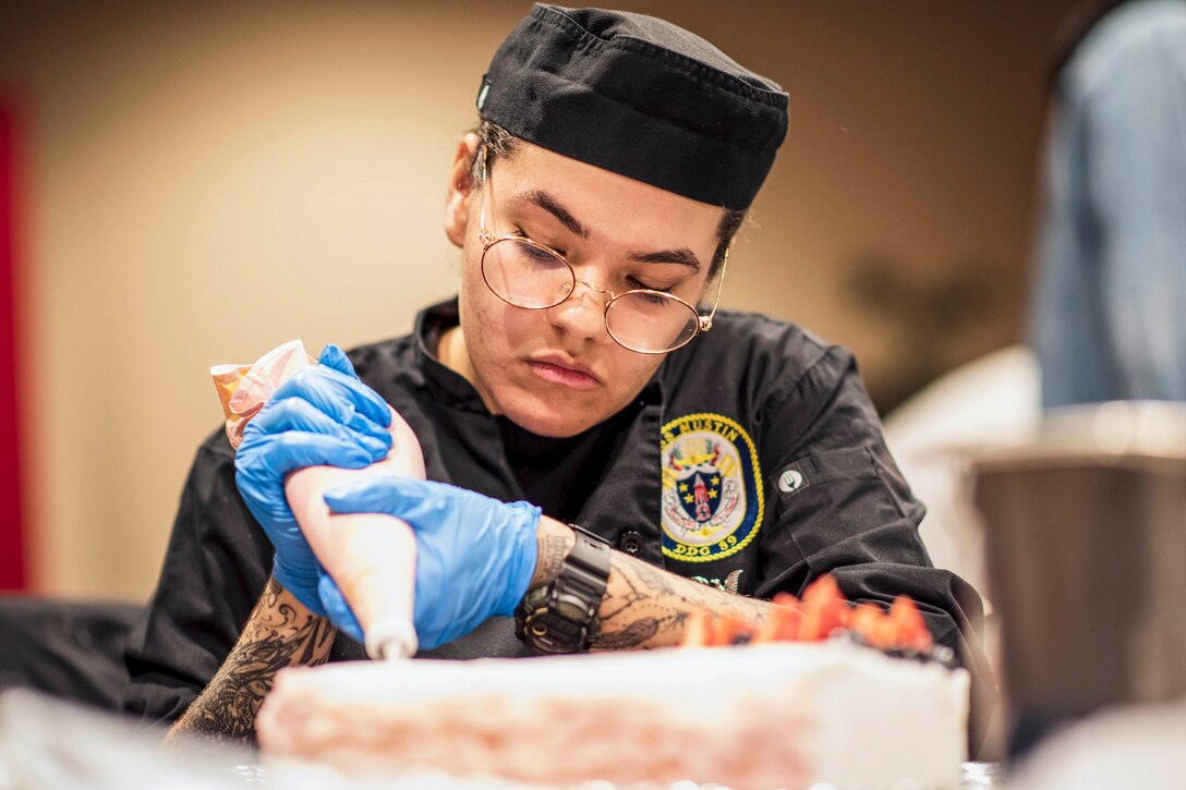 A sailor wearing light blue gloves uses a pastry bag to decorate a cake.