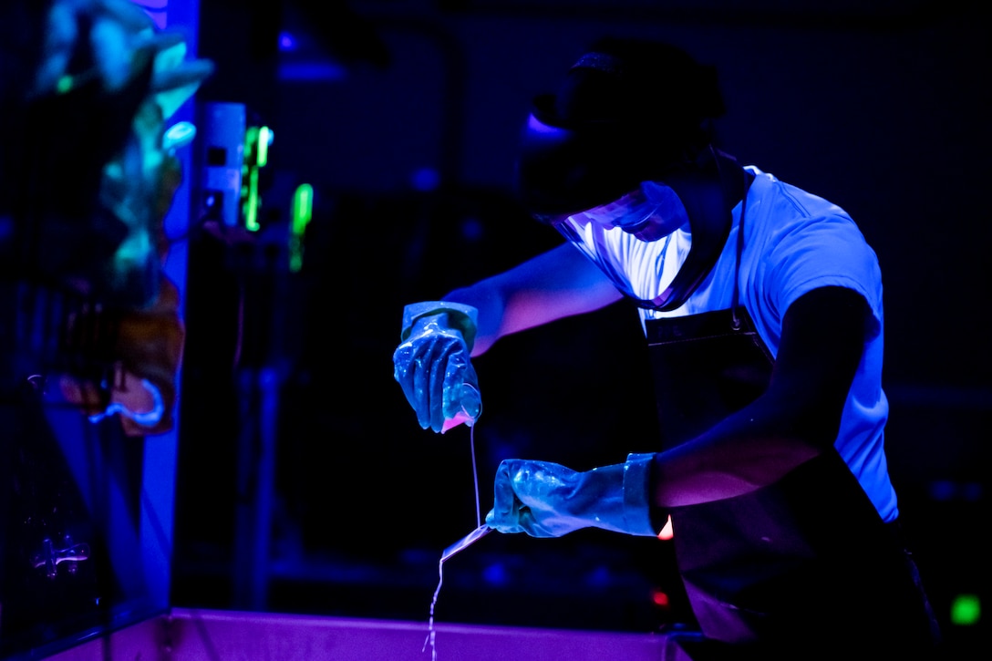 An airman wearing gloves and protective headgear works over a basin in a room illuminated by ultraviolet light.