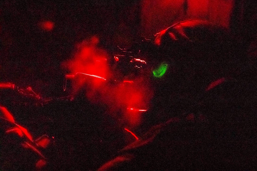 A Marine illuminated by red light looks through a rifle’s scope.