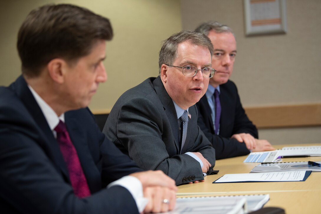 Man speaks while seated at a table between two other men.
