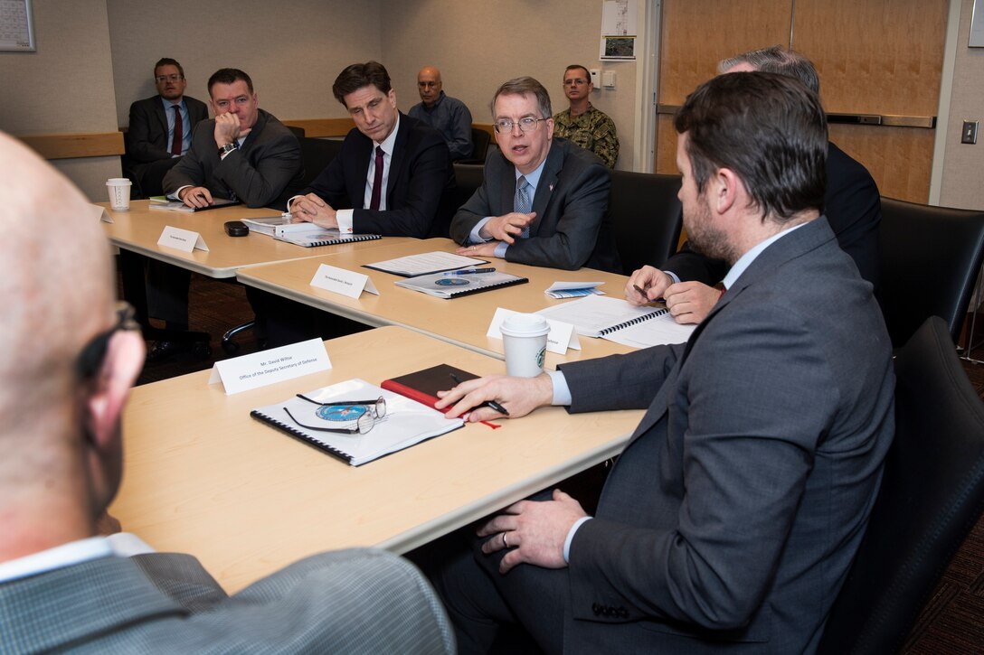 Man speaks to other men. All are seated around a table.