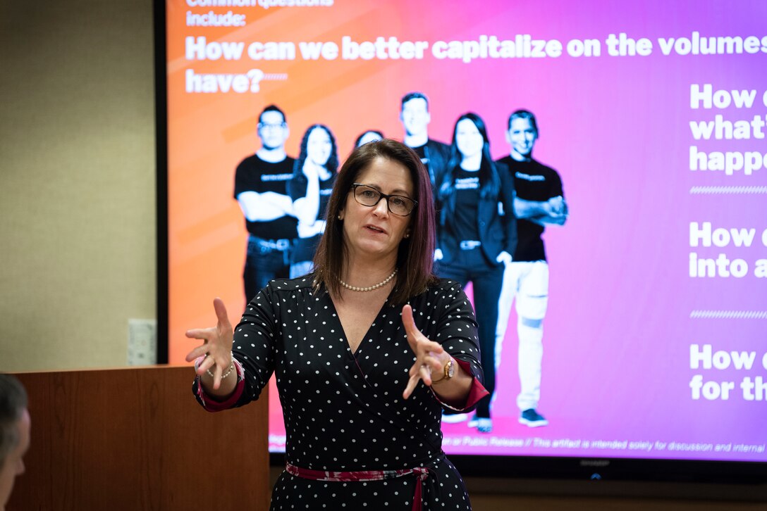 Woman standing in front of an illustrated briefing slide gestures with both hands while speaking.
