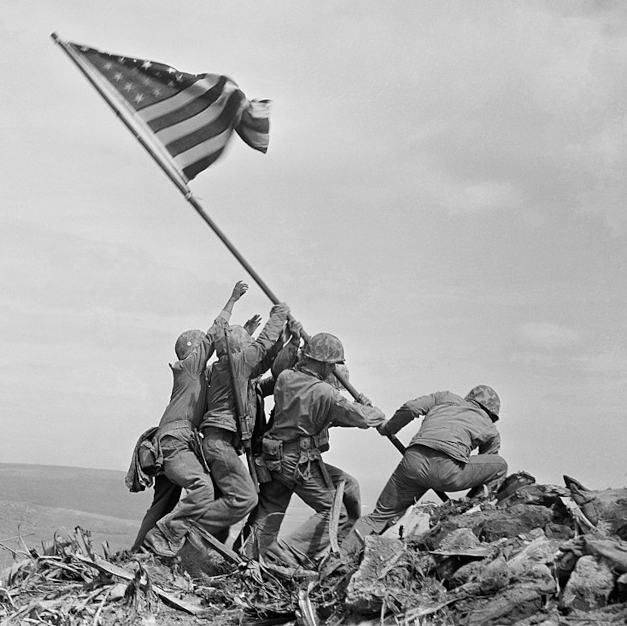 Black and white photo of Marines lifting an American flag to plant it on top of a mountain.