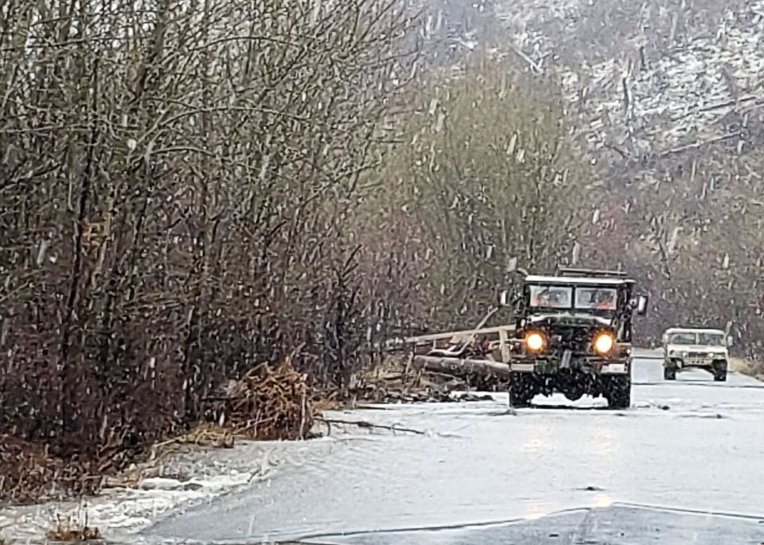 Two former military trucks drive on a road with flood water covering it.