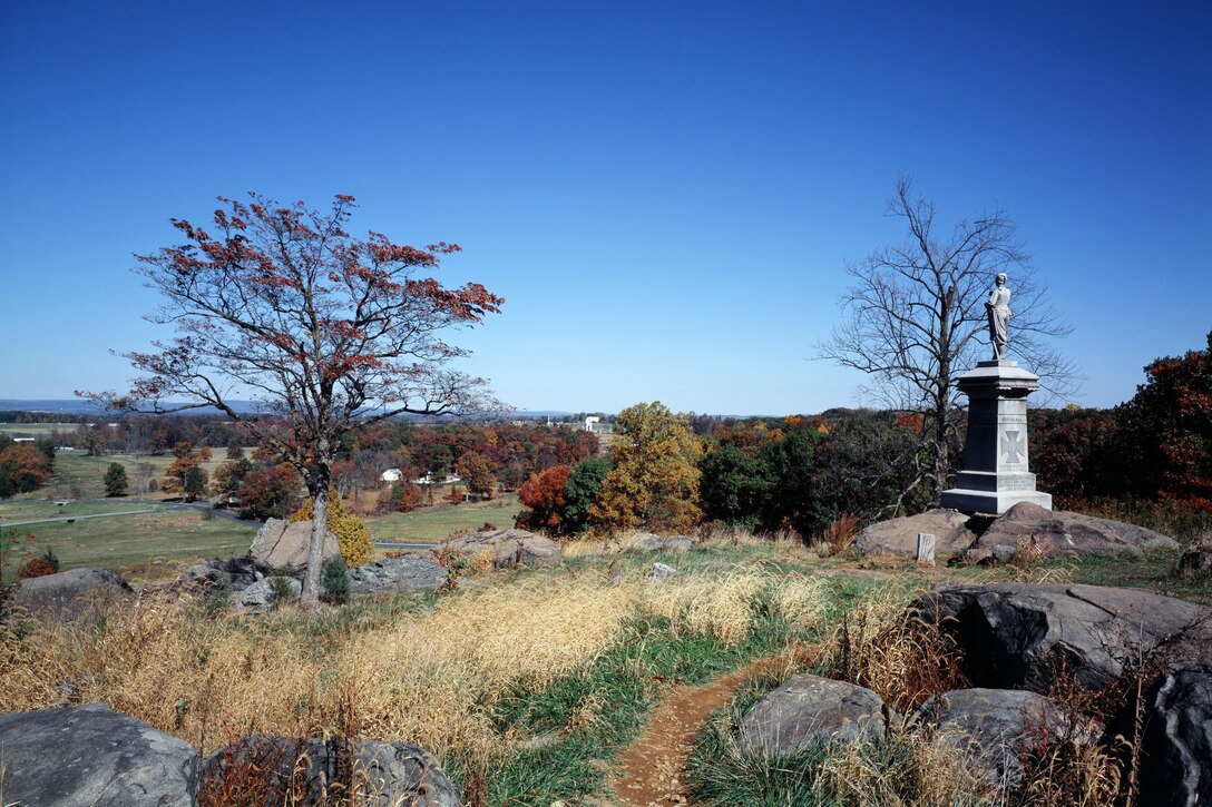 Rocks, autumn trees and brush cover a hillside. On the right is a monument with a man’s statue on top.