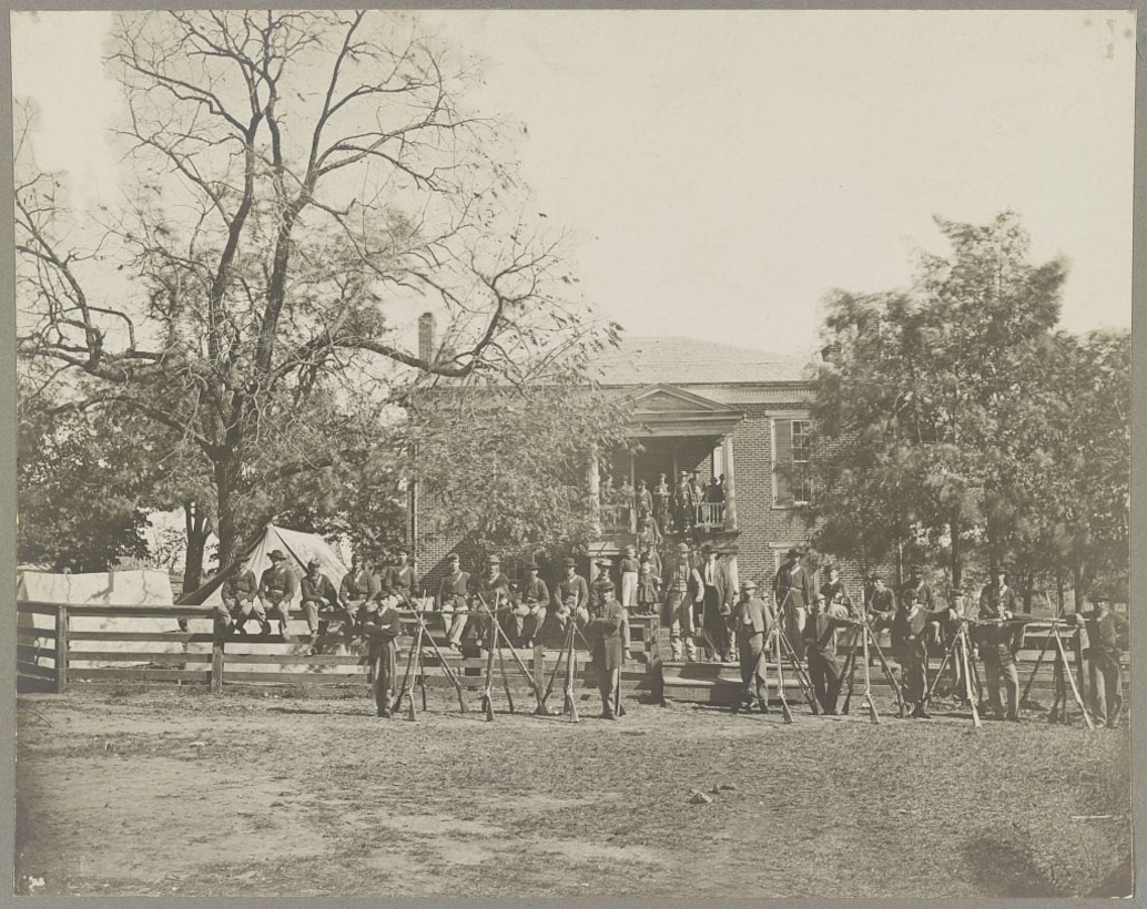 Dozens of Civil War soldiers holding muskets stand in front of a two-story building. More men sit on a fence, while others stand on a second-story balcony.