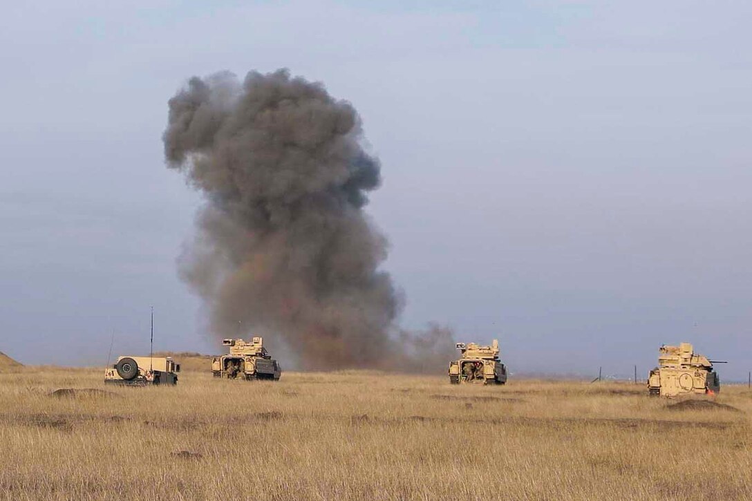 Military vehicles move through a field with a plume of smoke in the background.