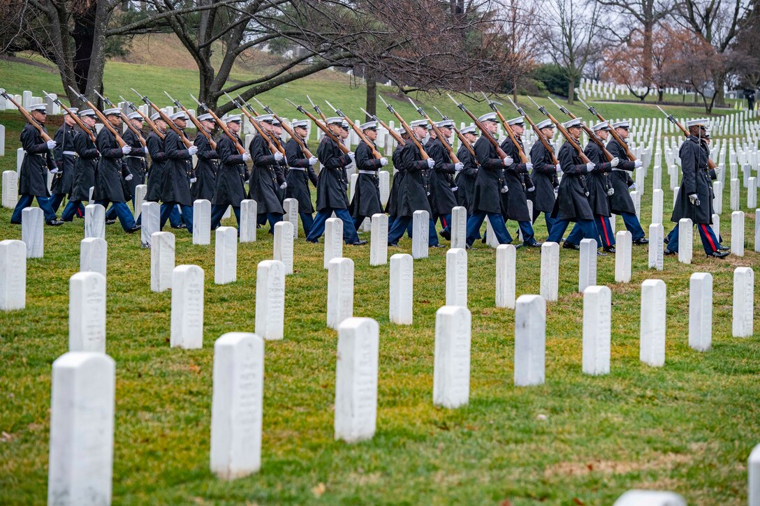 Soldiers and Marines walk in formation past gravestones in a cemetery.