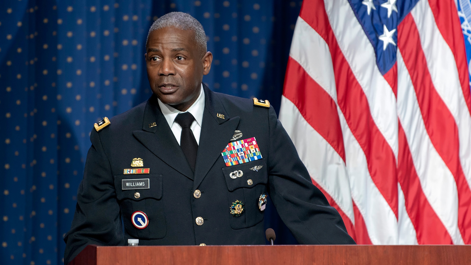 African-American man in Army dress uniform stands at a podium in front of the U.S. flag.