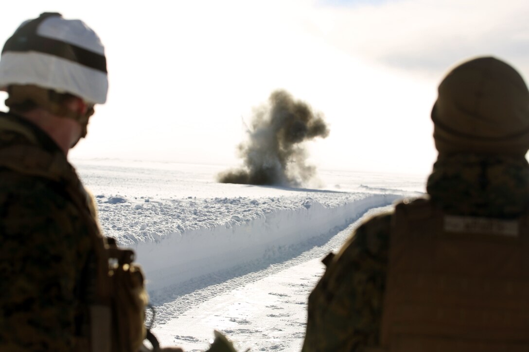 A cloud blooms out of a snowfield. Two service members watch.