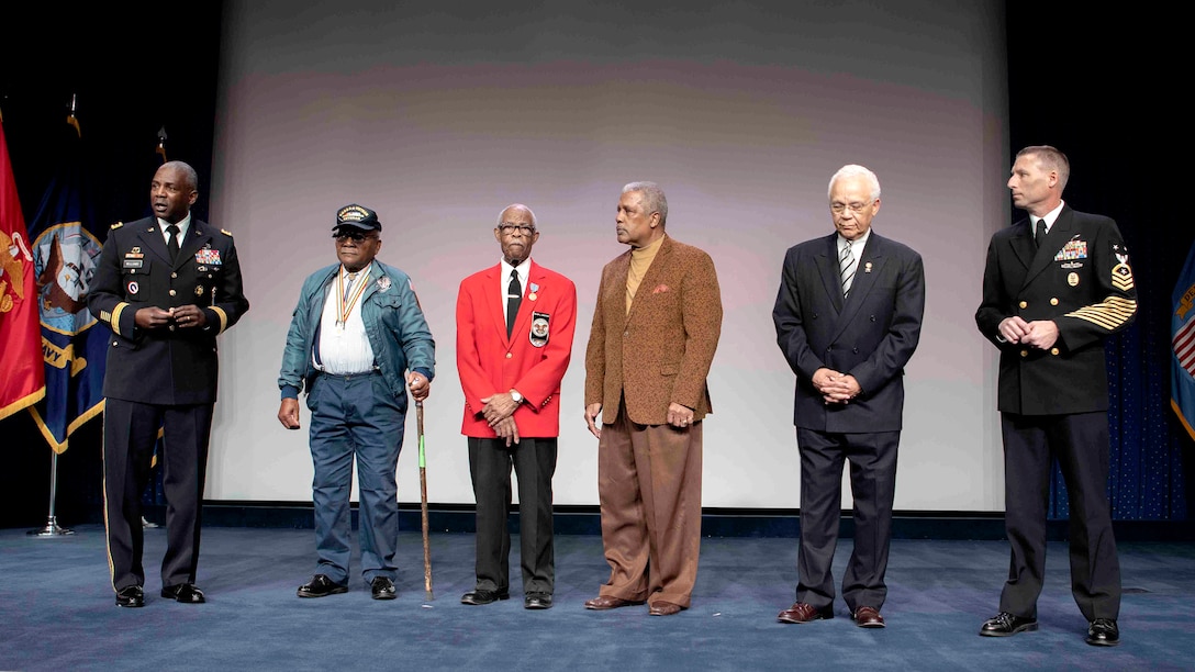 Six men stand on a stage. Man on the far left and far right are in military dress uniform.