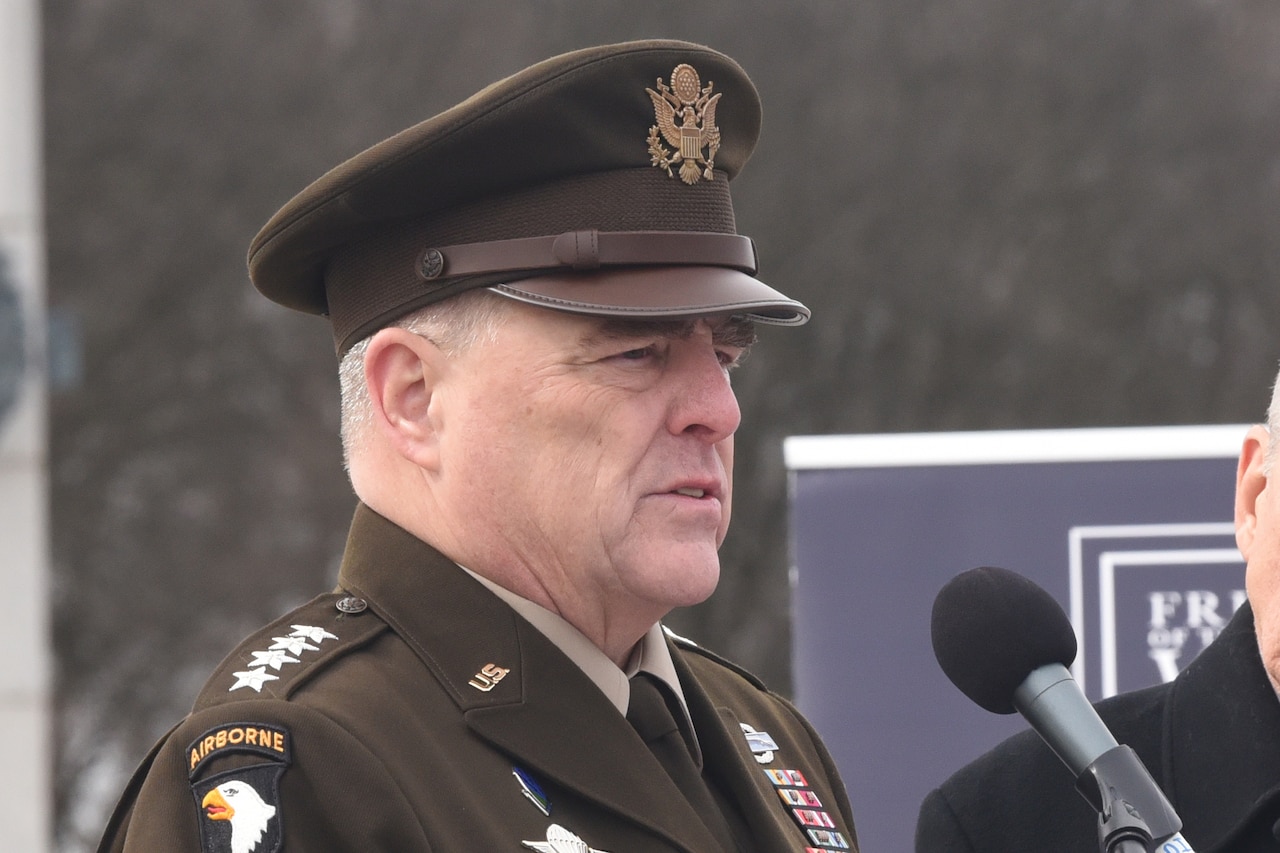 Four-star general speaks at a lectern as a World War II veteran listens in the foreground.