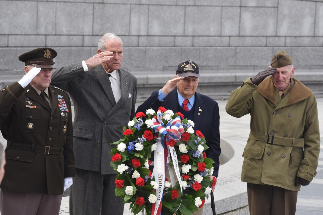 Four men, one in uniform, salute as they stand behind a wreath.