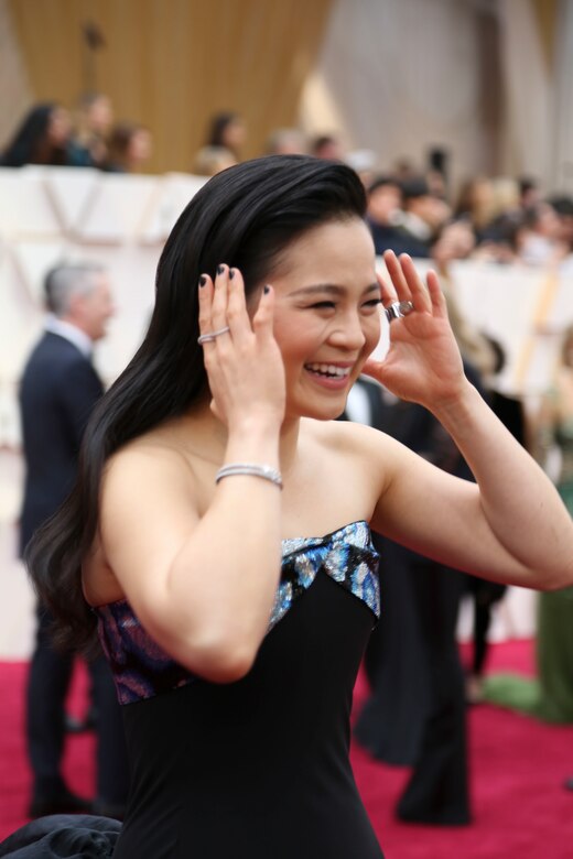 A smiling woman in a black dress holds her hands up to her head as if she’s about to push her hair back.