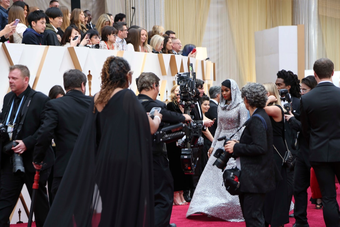 Photographers on a red carpet swarm a woman wearing a shimmering long silver gown with a matching hood on her head. Onlookers in stands above the carpet look down at her.
