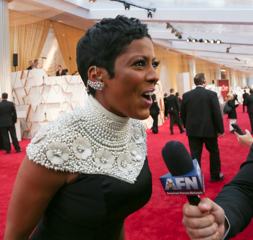 A woman in a black dress with pearl-beaded neckline talks to someone off-camera who’s holding a microphone on a red carpet.