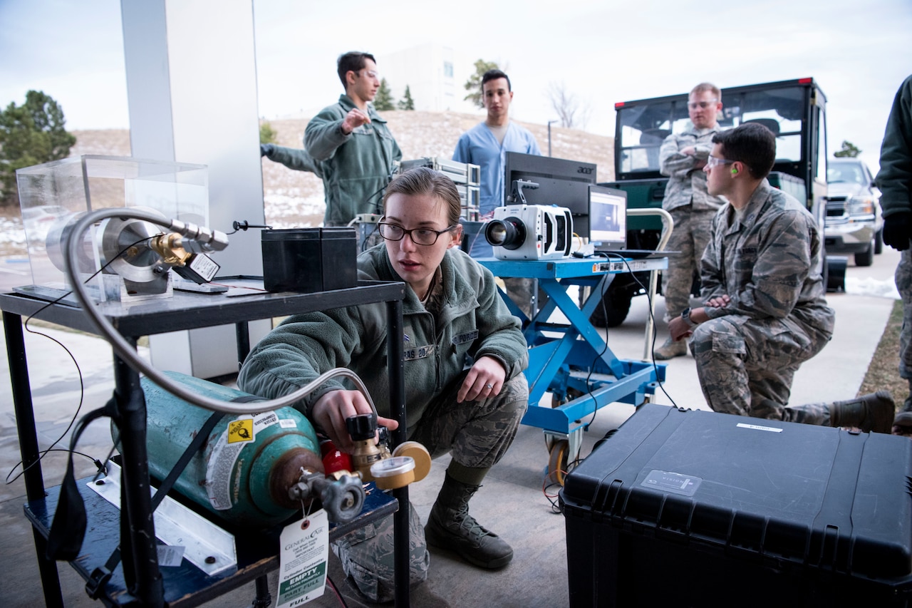 Military personnel work outdoors with equipment connected to a tiny jet engine.