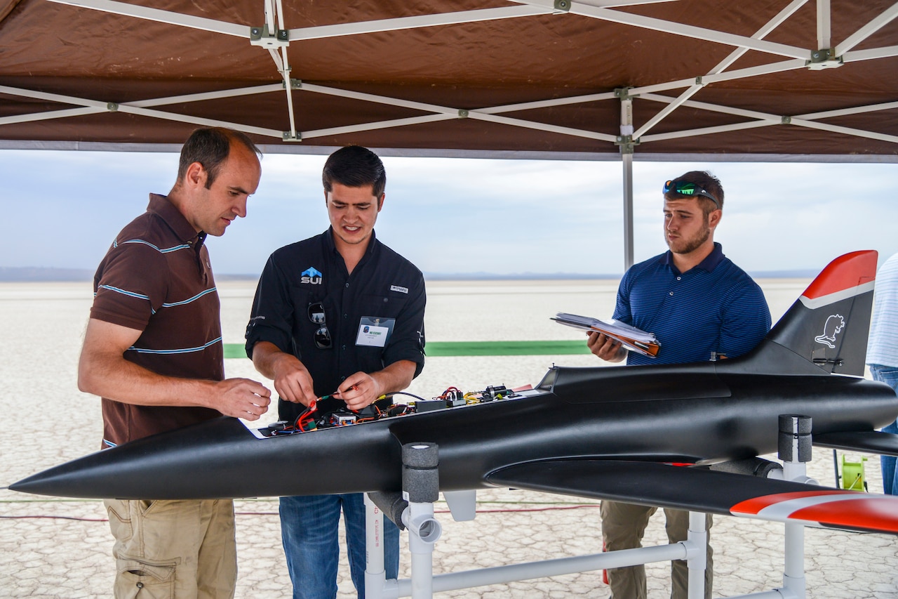 Three men stand under an overhang and work on a small aircraft.