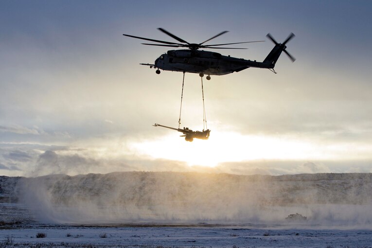 A helicopter, shown in silhouette, transports a howitzer hanging beneath it on slings.