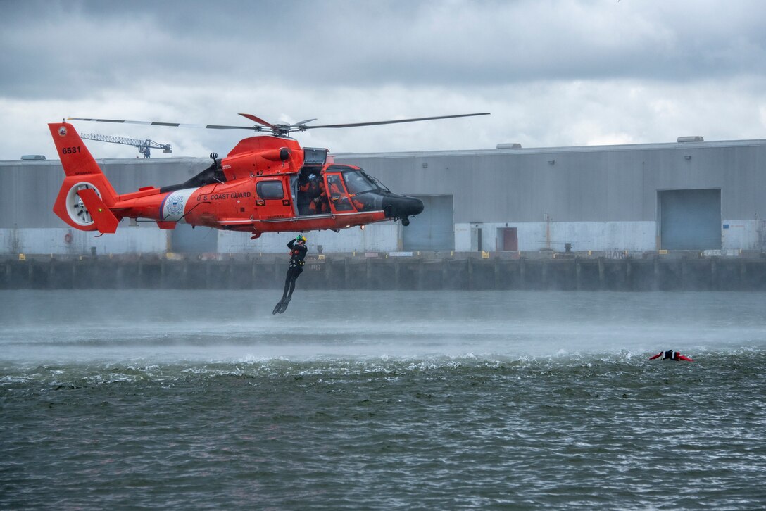 A Coast Guard member in a wetsuit jumps from an orange helicopter hovering over water.