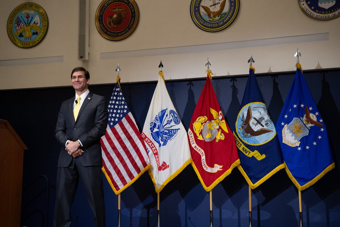 Defense Secretary Dr. Mark T. Esper stands on a stage with flags behind him.