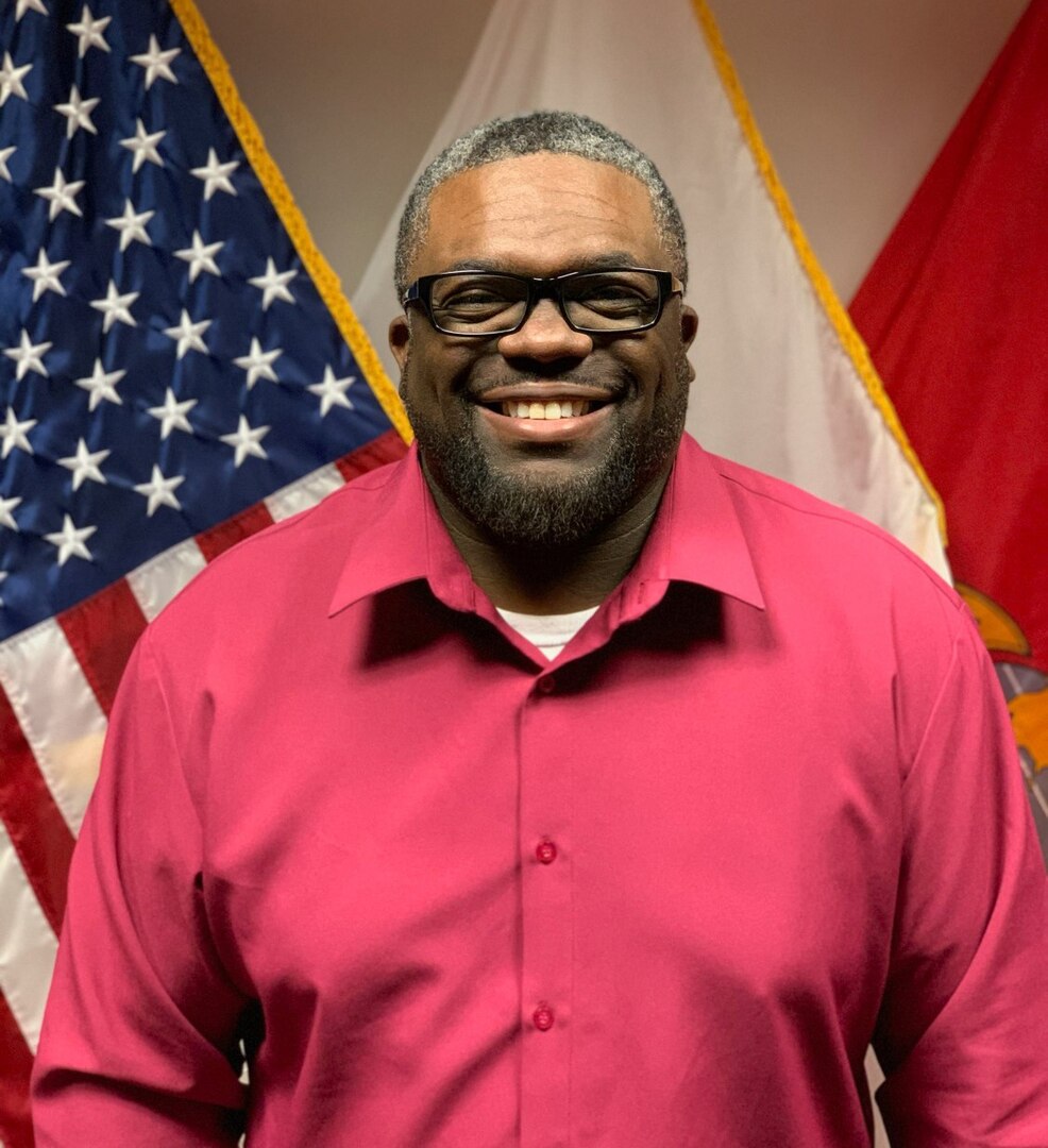 Photo depicts Bryant Daniels standing in front of a flag wearing a red shirt and glasses.