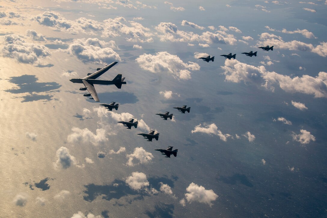 Eleven aircraft fly in a "V" formation over water in a cloud-studded sky.