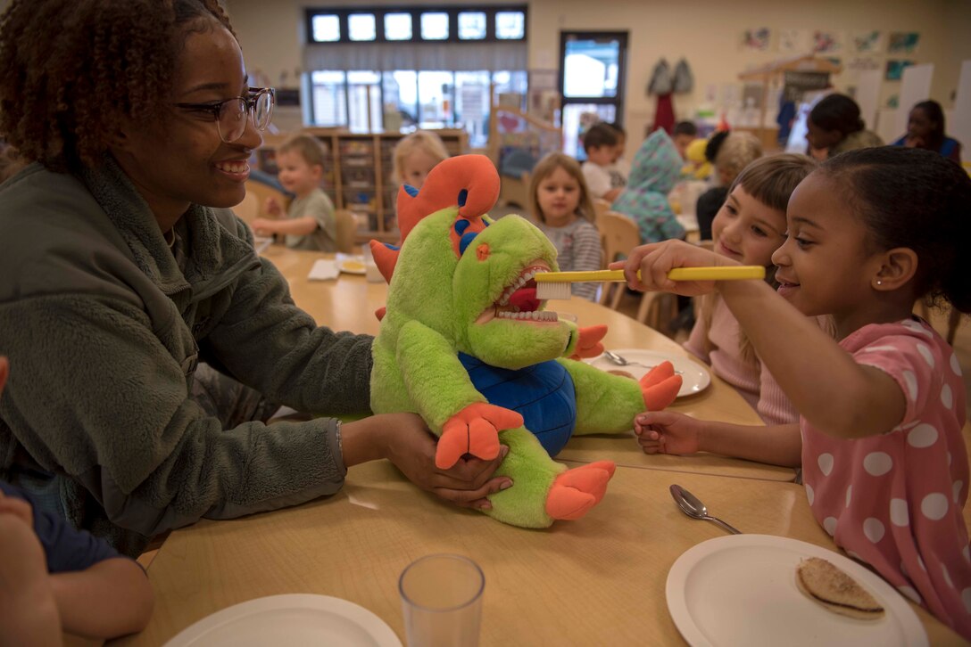 An airman watches a child brush a stuffed animal’s teeth while other children observe.