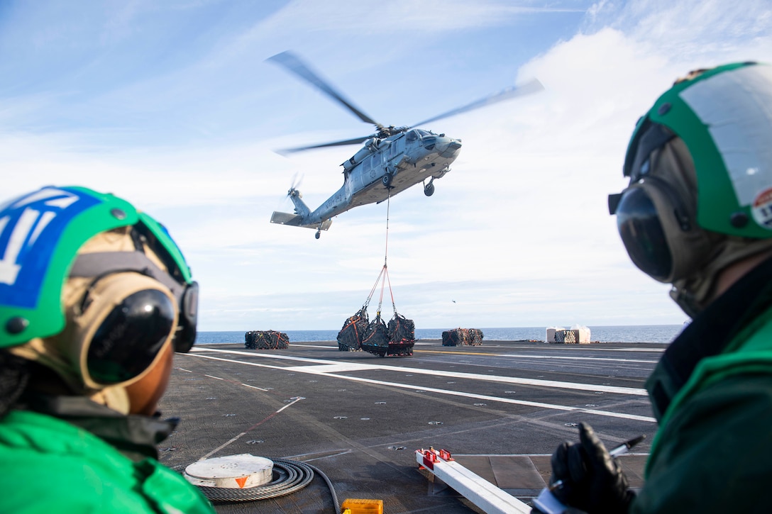 Sailors on a ship deck watch a hovering helicopter with cargo hanging from a sling.