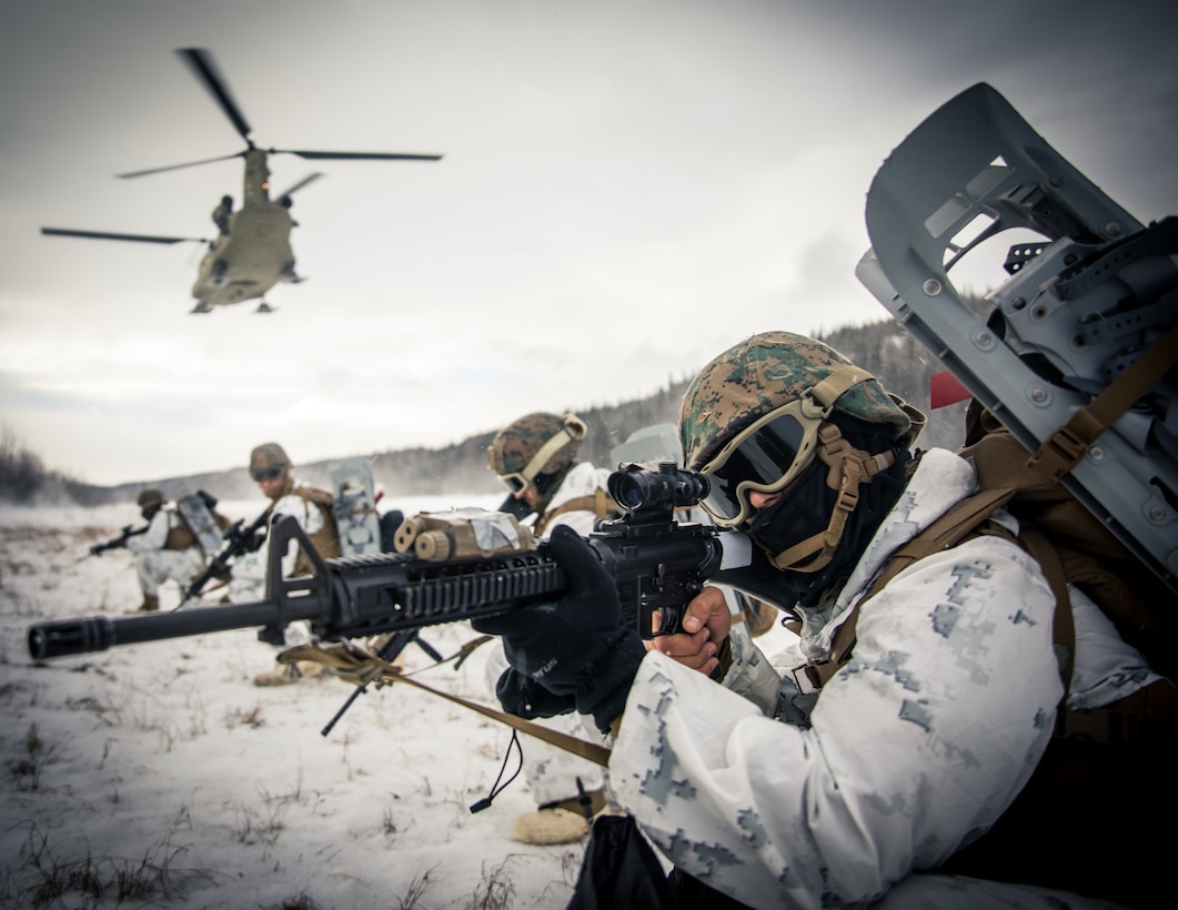 Marines point weapons in the snow while a helicopter passes by.