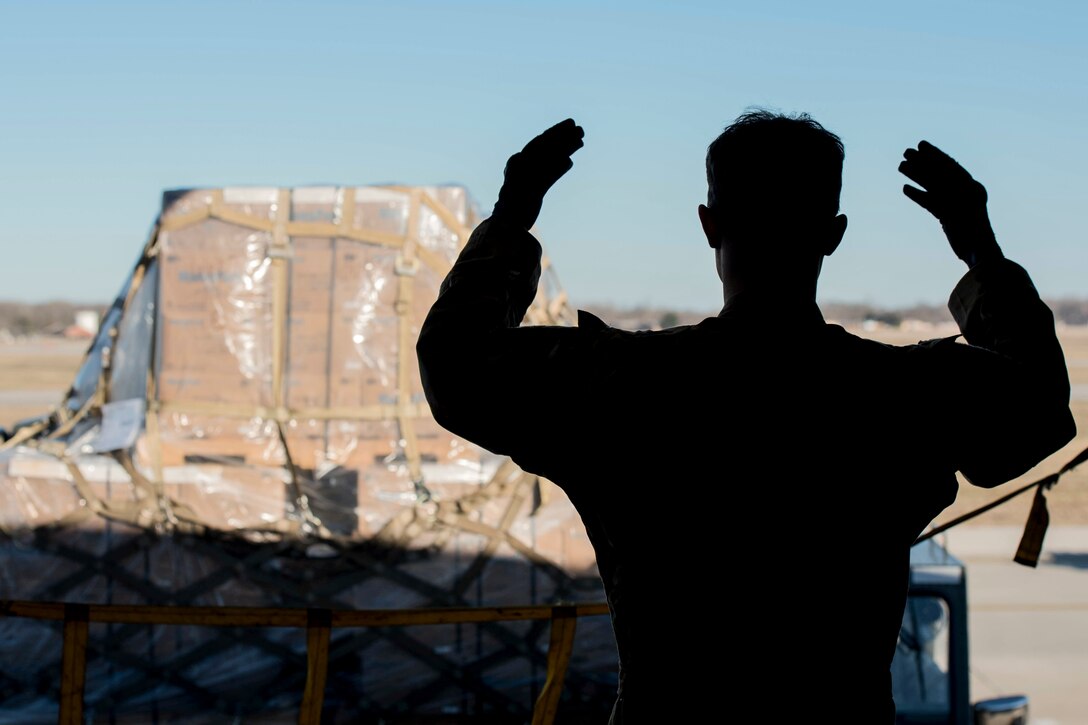An airman, shown from behind in silhouette, gestures with both arms in front of a vehicle carrying boxes.