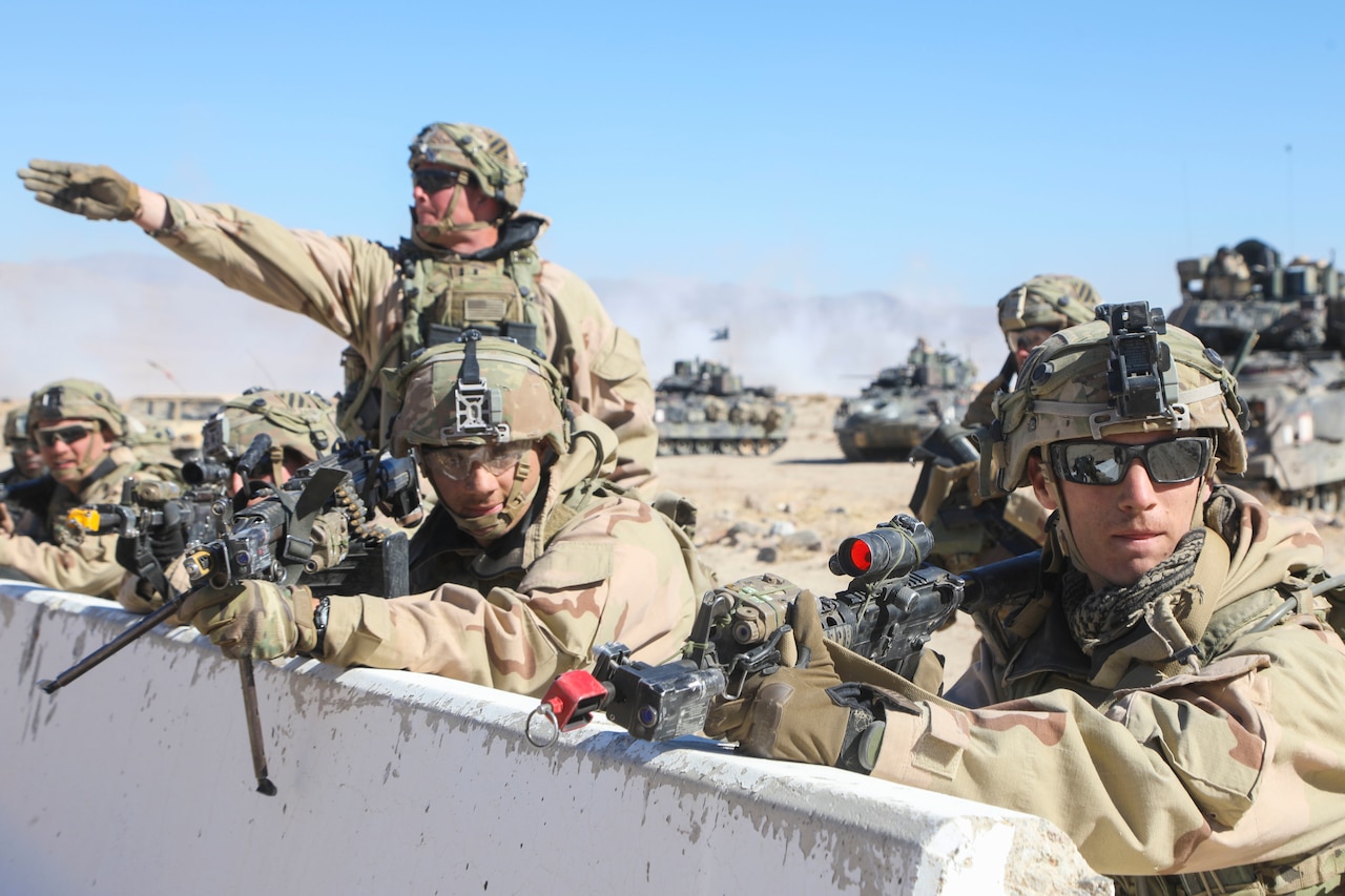 A soldier stands and gestures forward with one arm, while others hold up weapons from behind a barrier.