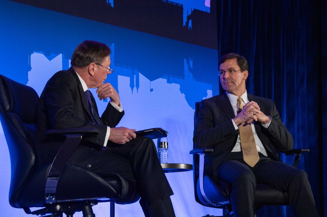 Defense Secretary Dr. Mark T. Esper speaks to another man while seated on a stage.