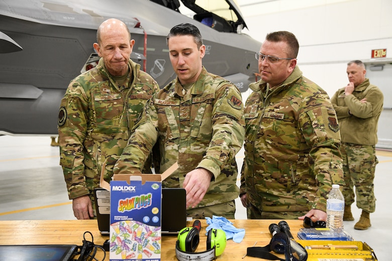 Tech. Sgt. Matthew Ruggiero (middle), 4th Aircraft Maintenance Unit, Gen. Mike Holmes, commander of Air Combat Command, left, and  Command Chief Master Sgt. David W. Wade, command chief of ACC, look over equipment on a table located in a hangar at Hill AFB. The nose of an F-35A Lightning II can be seen in the background.