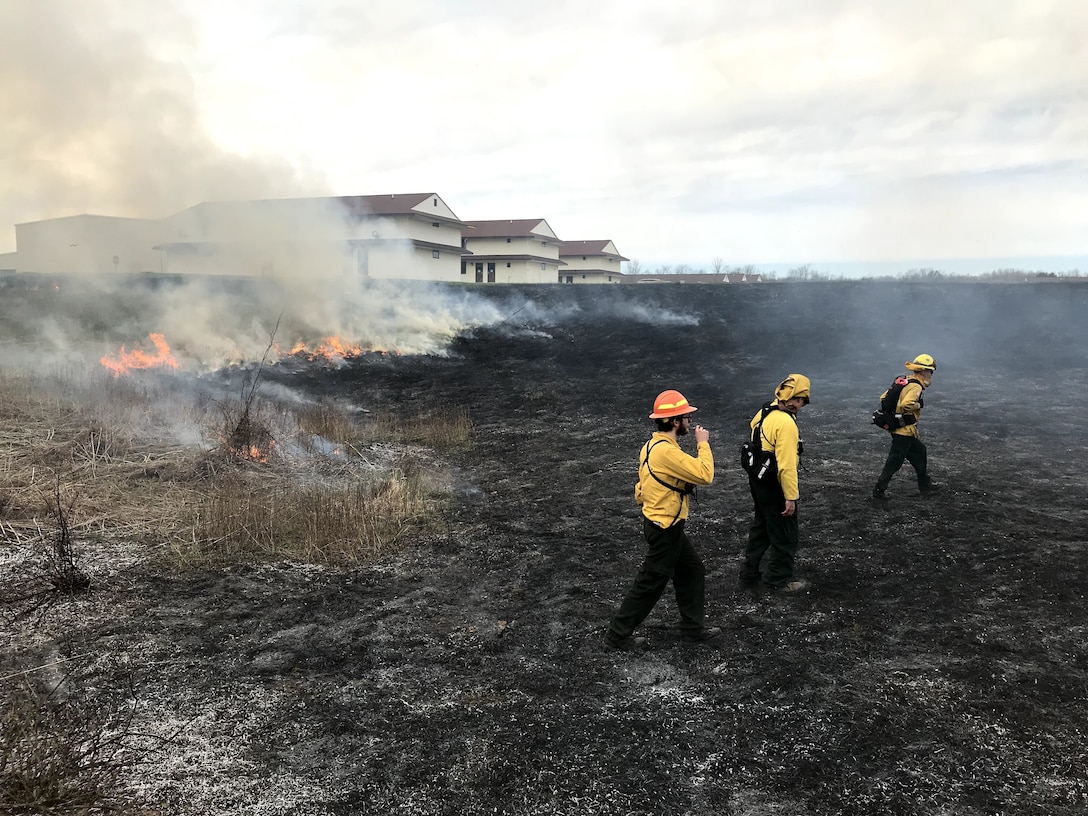 People in yellow shirts and hard hats walk away from a fire line on a smoldering field.