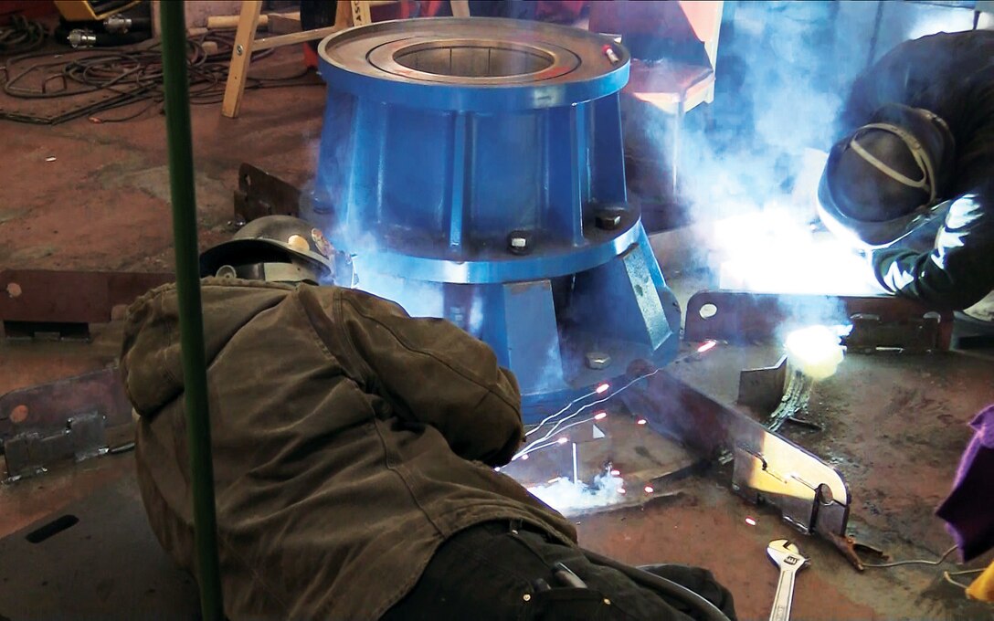 The Dredge Essayons receives an entirely new steering system that requires cutting holes on both sides of the ship. Inside the ship, two workers weld in the port-side rudder bearing assembly