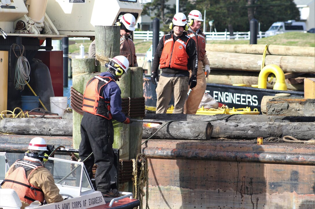 A five-person crew wearing life vests use a torch to deconstruct wooden pilings